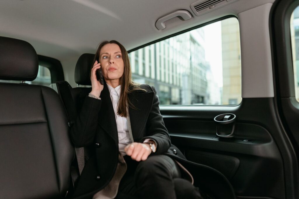 A professional woman in a blazer talks on her smartphone while seated in a car.
