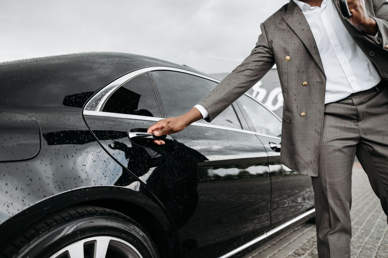 Home A man in a suit opens a luxury car door under raindrops on stone pavement.